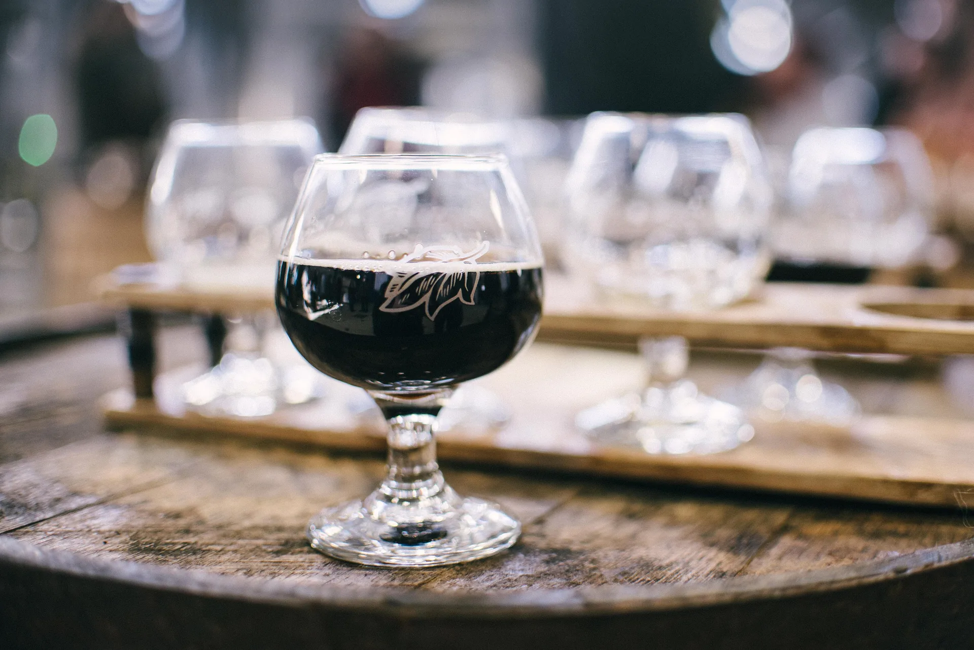 Copo de cerveja escura sobre um barril de madeira, com o bar desfocado ao fundo